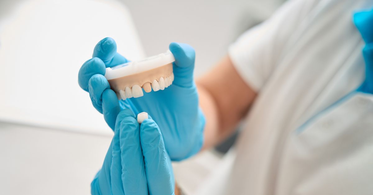 A dental professional wearing blue gloves holding a prosthetic tooth crown and a dental model.